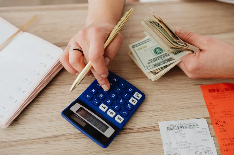 Hands using a calculator beside a stack of cash and receipts on a desk