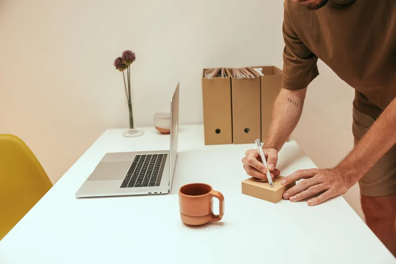 Person writing notes at a laptop with a coffee mug on a minimalist desk