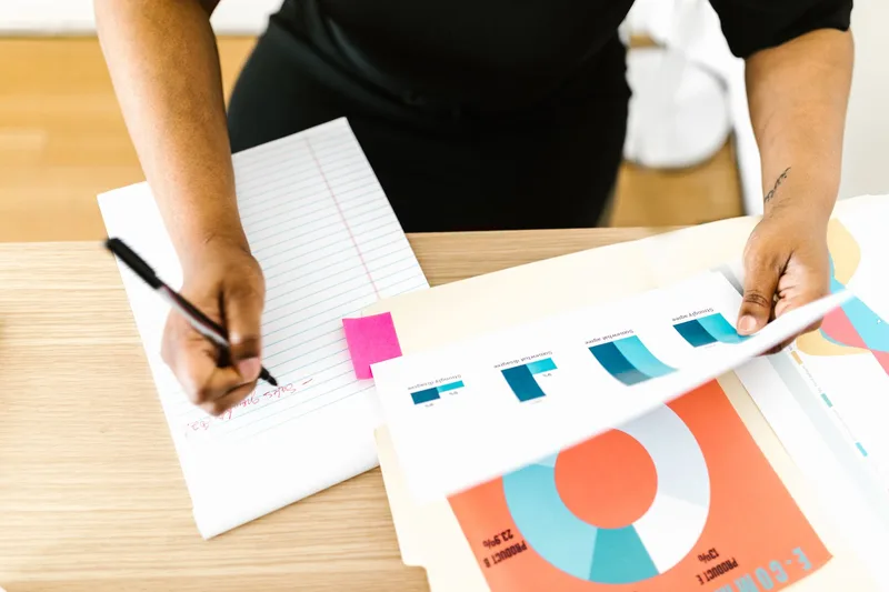 Person writing notes while reviewing printed charts and revenue reports at a desk