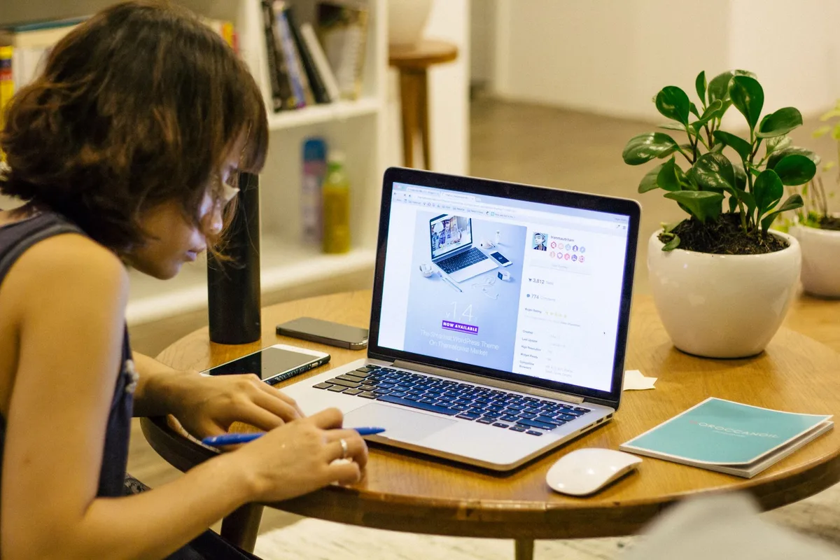 Woman writing in a notebook beside an open laptop on a wooden desk with a small plant