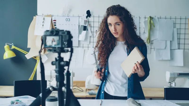 Woman holding a notebook and speaking to a camera on a tripod while recording a video lesson in her studio