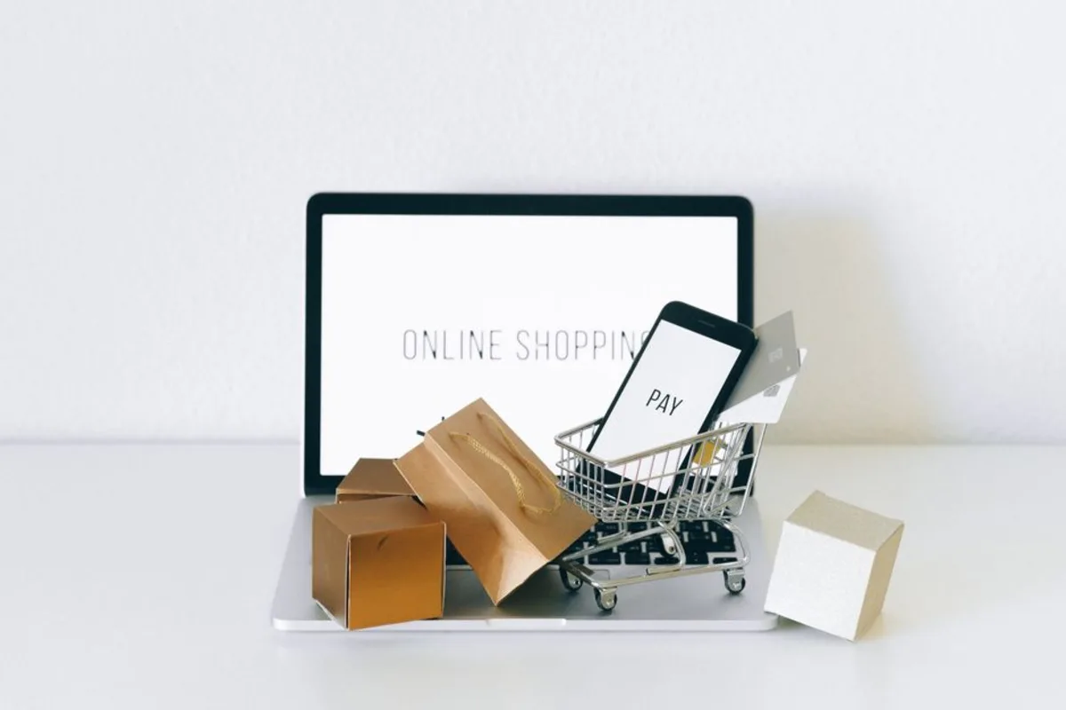 Laptop displaying “online shopping” text on screen with a miniature shopping cart, a phone showing a pay screen, and small packages on the keyboard